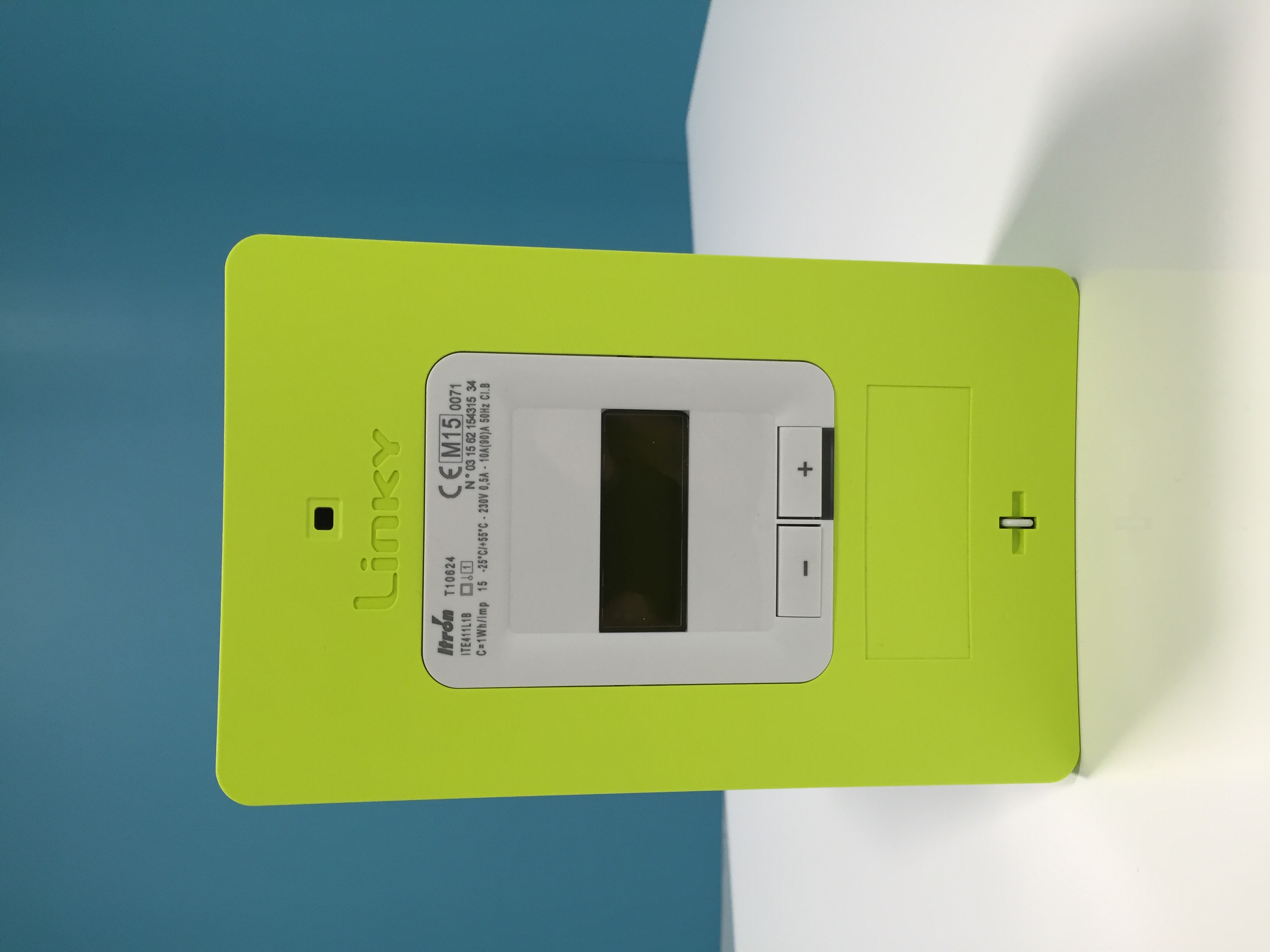 Compteur vert Linky posé, capots en place, deux boutons – et +, écran sombre ; marque Itron et 230 V visibles.